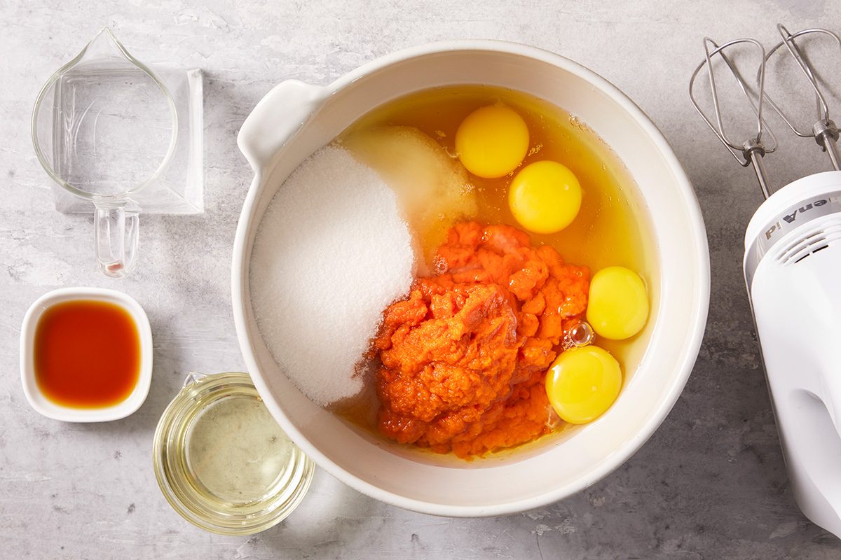 A mixing bowl with eggs, sugar, pumpkin puree, and other ingredients, surrounded by a measuring cup with water, a small bowl of vanilla extract, oil, and a hand mixer on a gray countertop.