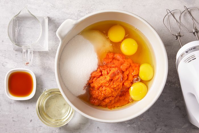 A mixing bowl with eggs, sugar, pumpkin puree, and other ingredients, surrounded by a measuring cup with water, a small bowl of vanilla extract, oil, and a hand mixer on a gray countertop.