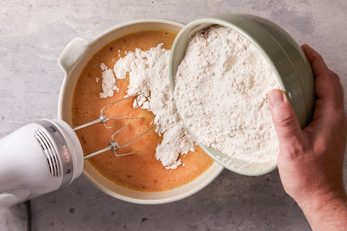 A hand pours flour from a bowl into a mixing bowl containing an orange batter while an electric hand mixer with beaters is in the bowl, ready to blend the ingredients together.