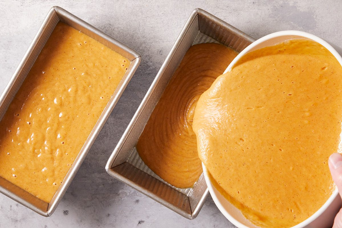 Two loaf pans on a counter, one filled with orange batter and the other being filled as someone pours more orange batter from a mixing bowl.