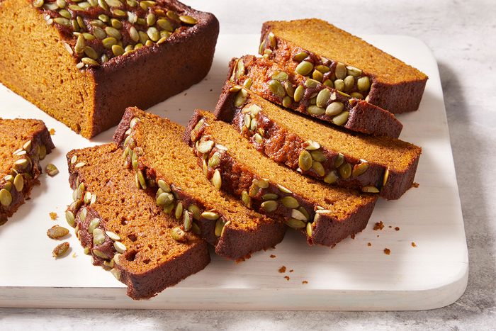 Sliced pumpkin bread topped with green pumpkin seeds, arranged on a white cutting board against a light background.