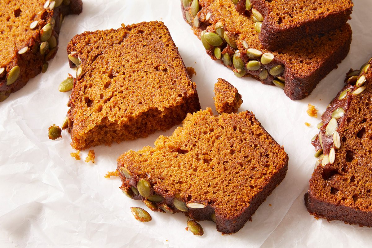 Sliced pumpkin bread with a browned crust and seeds on top, arranged on white parchment paper. One slice is broken into pieces, showing the moist texture inside.
