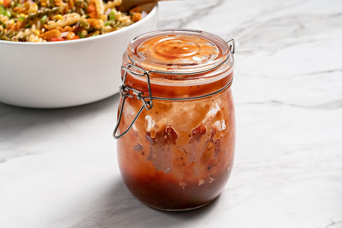 A glass jar filled with sweet and sour sauce sits on a white marble surface, with a bowl of stir-fry noodles blurred in the background.
