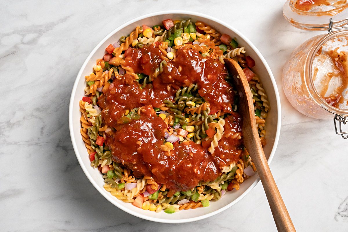 A white bowl filled with colorful rotini pasta topped with a thick, reddish sauce, with a wooden spoon on the side. Two glass jars with remnants of sauce are next to the bowl on a marble surface.