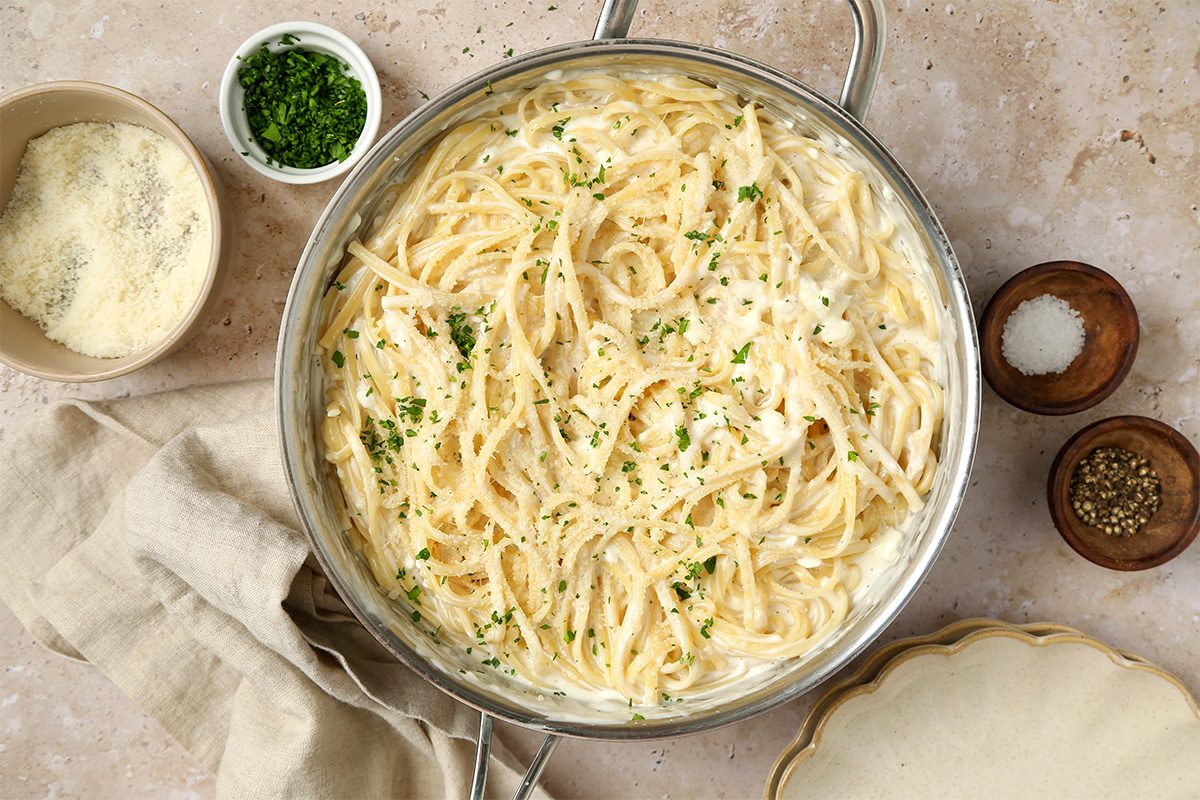 A pot of creamy spaghetti Alfredo topped with chopped parsley, surrounded by bowls of grated cheese, chopped herbs, salt, and pepper on a beige countertop with a beige napkin and an empty plate nearby.