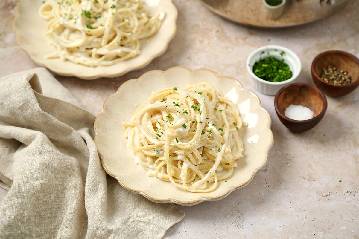 A plate of creamy pasta garnished with chopped herbs sits on a beige surface, with a napkin nearby and small bowls of salt, pepper, and chopped herbs in the background.