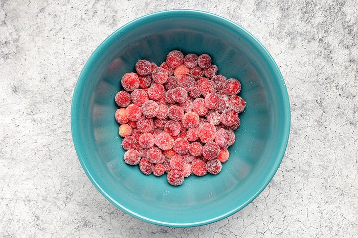 A teal bowl filled with frozen red cherries sits on a light gray, textured surface. The cherries are covered with a thin layer of frost.