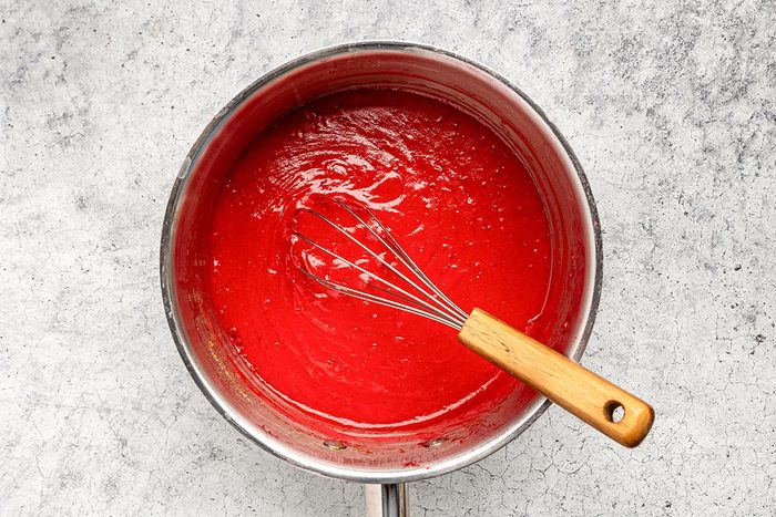 A metal saucepan filled with bright red sauce being whisked with a metal whisk that has a wooden handle, set on a light gray textured surface.