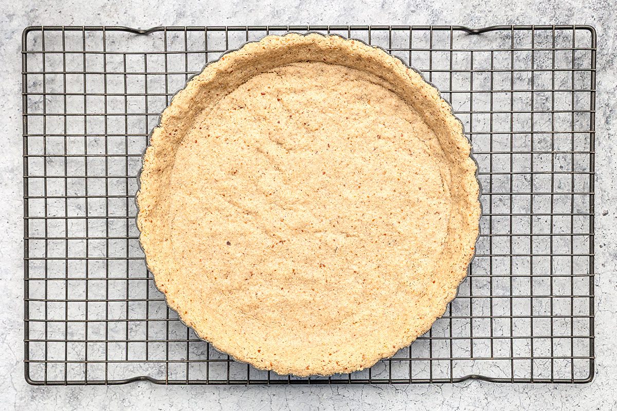 A baked tart crust sits on a metal cooling rack, viewed from above on a light gray textured surface. The crust is golden brown and evenly baked, with scalloped edges.