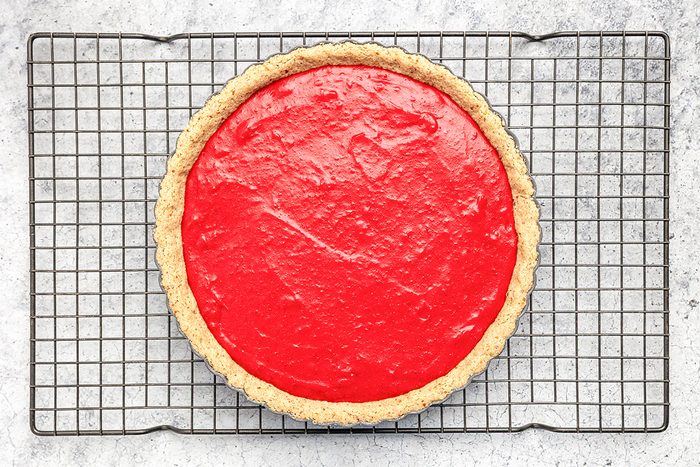 A round pie with a bright red filling sits on a cooling rack over a light gray, textured surface. The pie has a golden, crumbly crust and a smooth, glossy top.