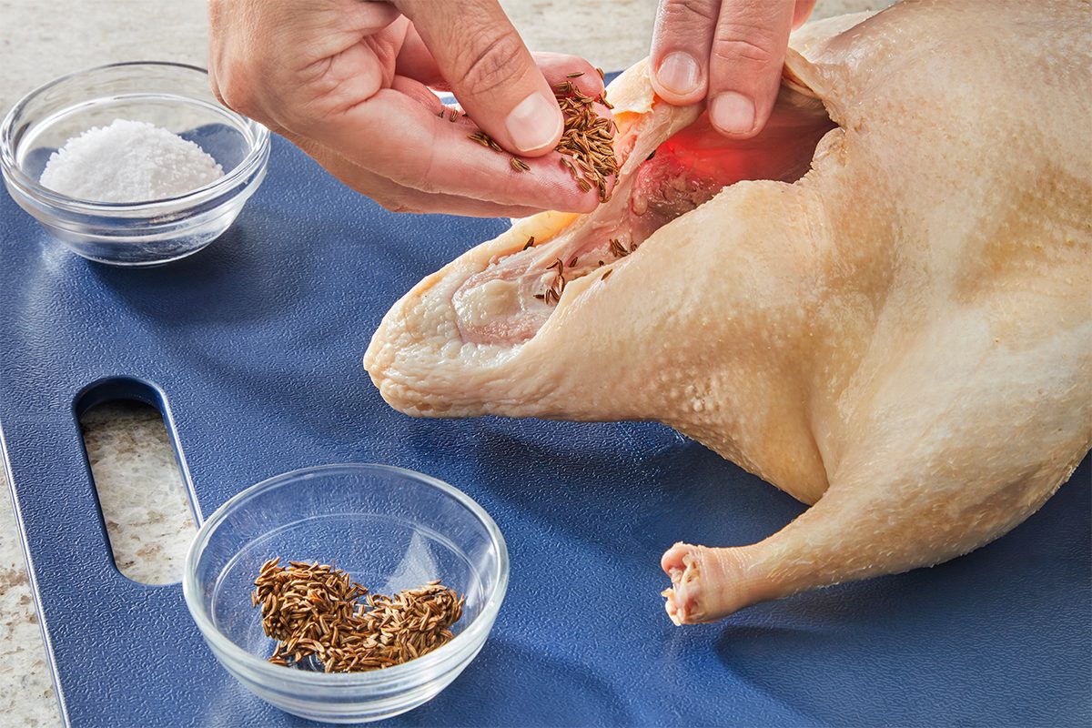 A person stuffs spices into the cavity of a raw whole chicken on a blue cutting board. Two small bowls, one with salt and one with seeds, are nearby.