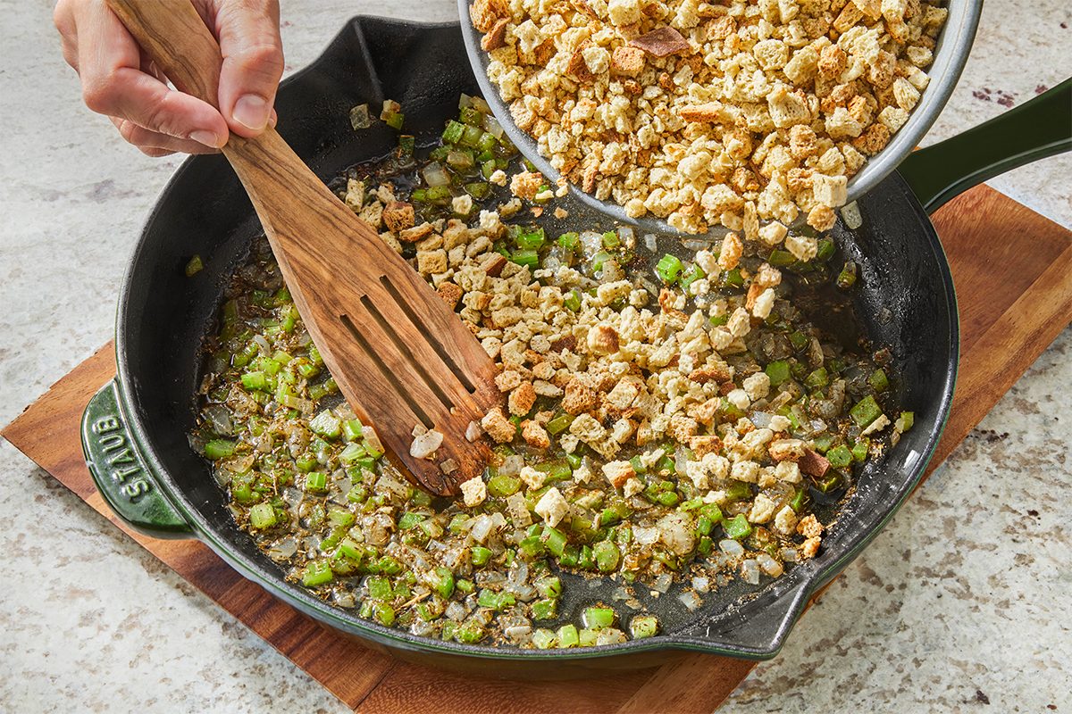A person stirs chopped celery and onions in a cast iron skillet while pouring in a bowl of bread cubes, preparing stuffing on a wooden board.