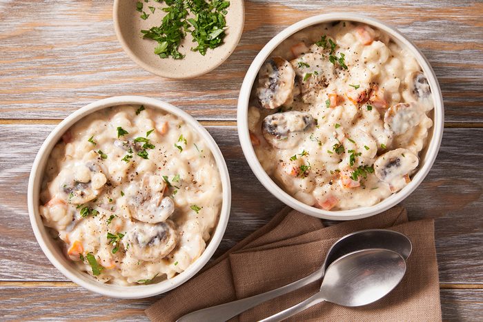 Two bowls of creamy mushroom and rice soup garnished with chopped parsley, placed on a wooden surface with a small dish of herbs and two spoons on a napkin nearby.