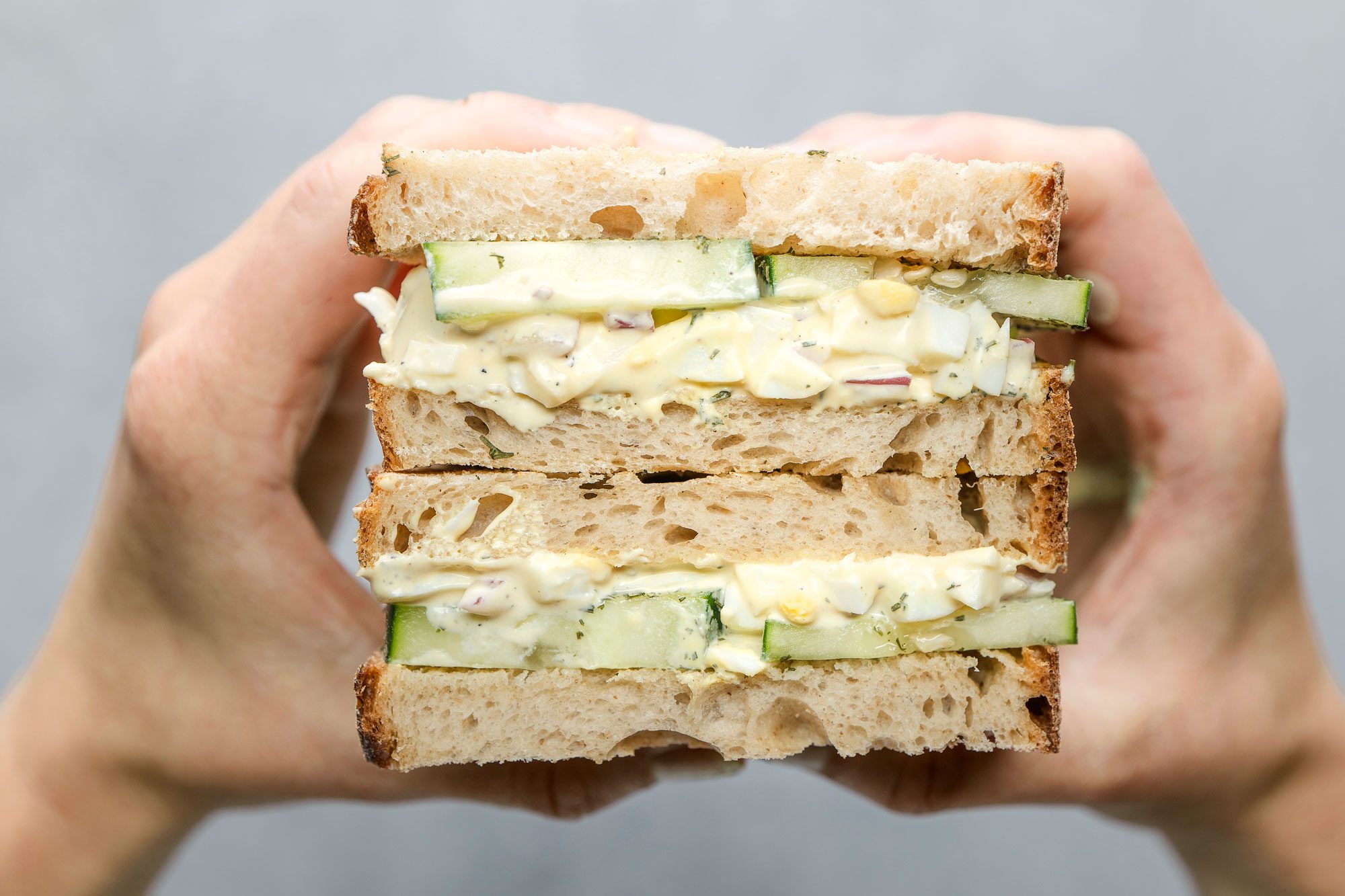 Close-up shot of Egg Salad and Cucumber Sandwiches held in both hands, showing layers of creamy egg salad and cucumber between slices of rustic bread