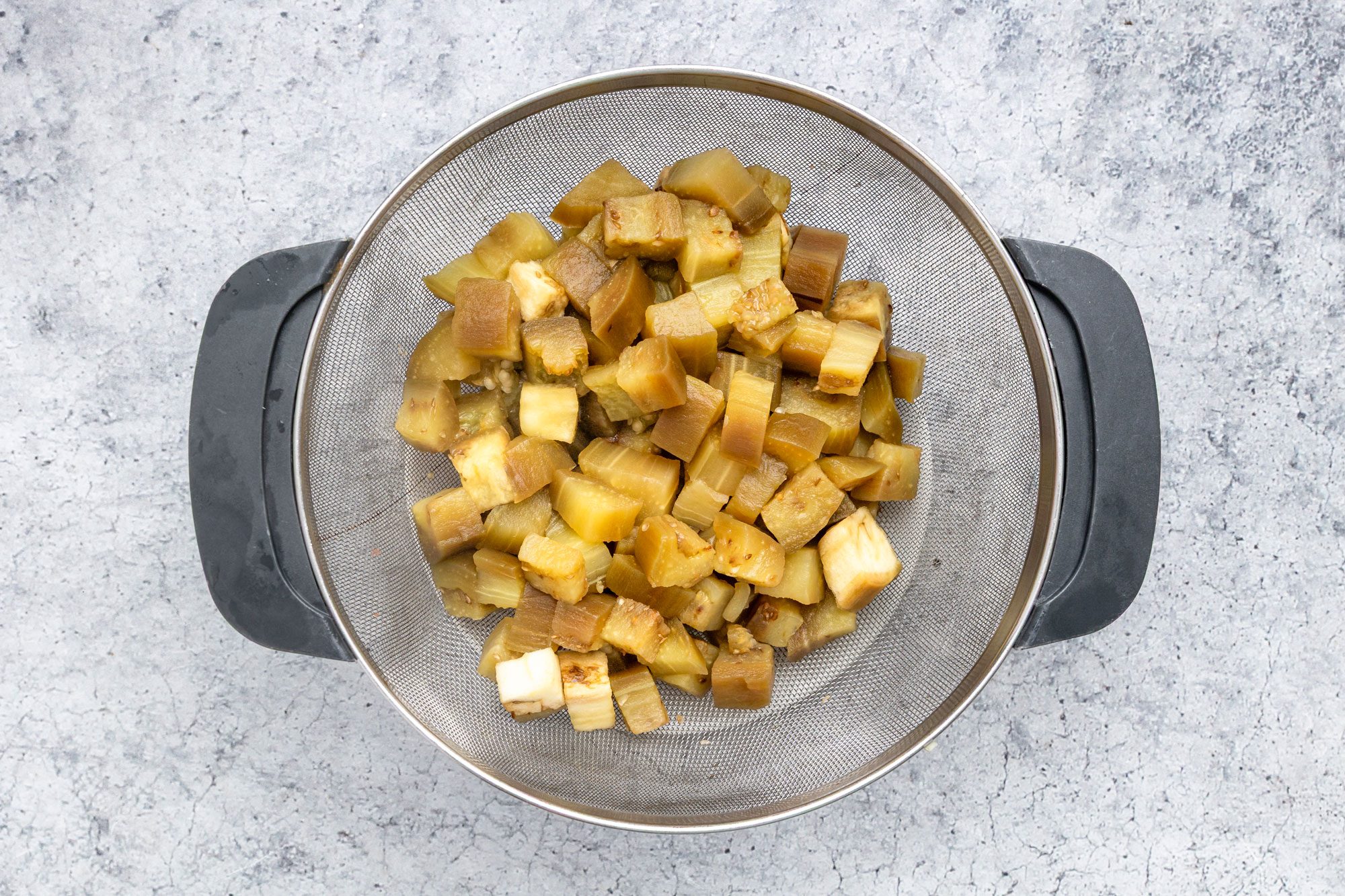 overhead shot of Diced pieces of cooked eggplant are draining in a metal strainer with black handles, placed on a gray textured surface