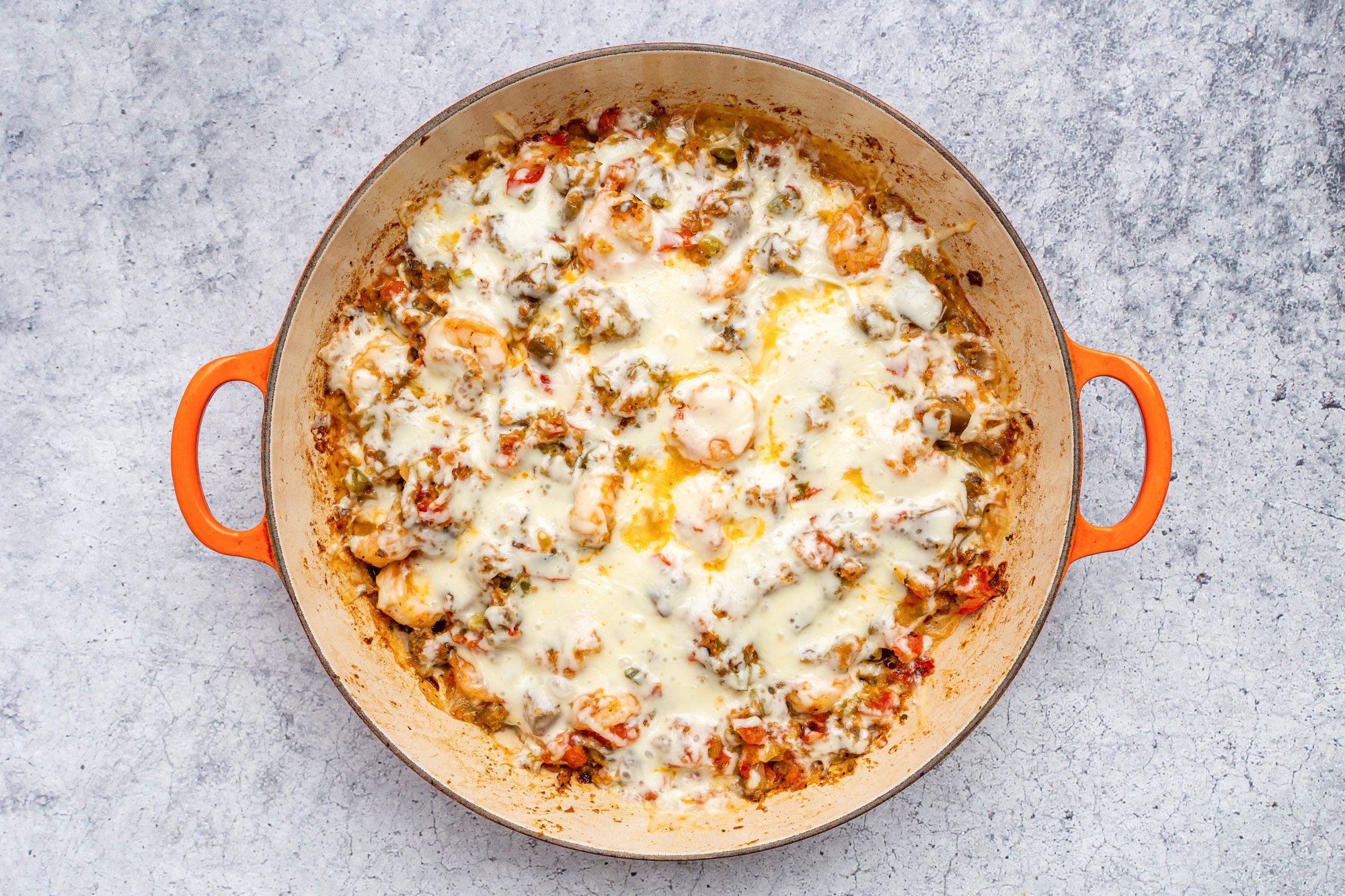 overhead shot of a round orange handled skillet filled with a baked dish featuring melted cheese, vegetables, and sauce on a light gray textured surface