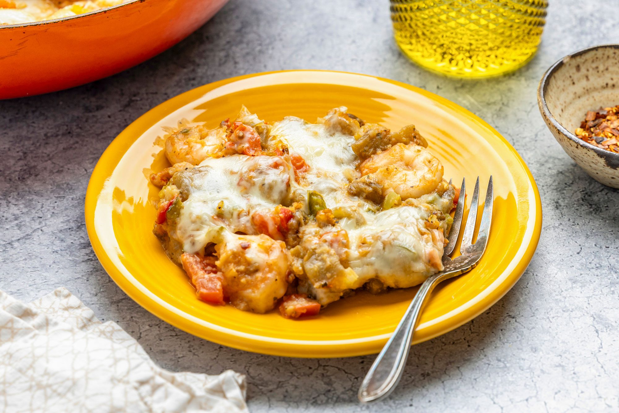 3/4th shot of ayellow plate with a serving of cheesy shrimp casserole, featuring shrimp, melted cheese, and vegetables, with a fork on the side, A bowl of the casserole, crushed red pepper, and olive oil are nearby