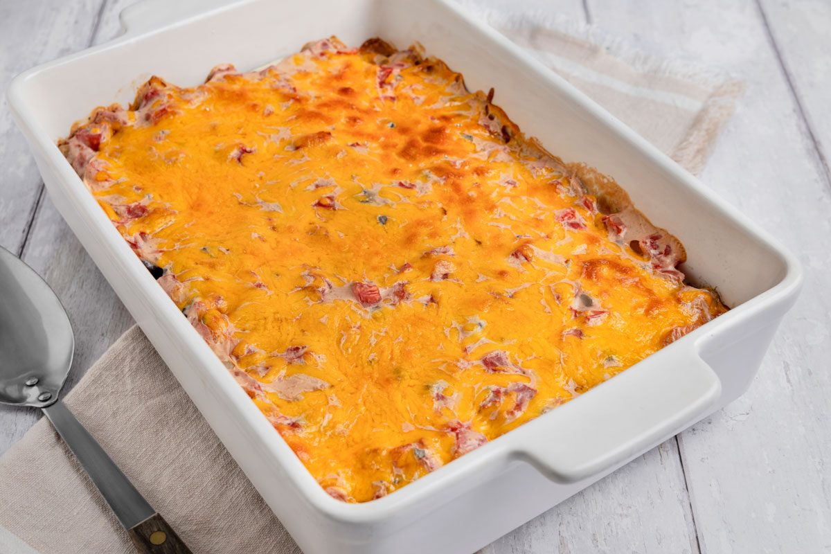 3/4 angle view shot of Firecracker Casserole in a baking dish with a serving spoon over a cream napkin; all set on a white wooden surface;