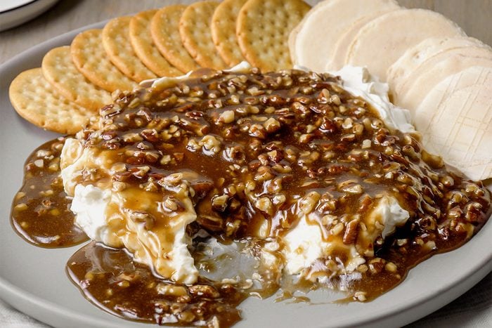A plate with round crackers and creamy cheese topped with a thick brown pecan and caramel sauce, partially spread, ready for serving.