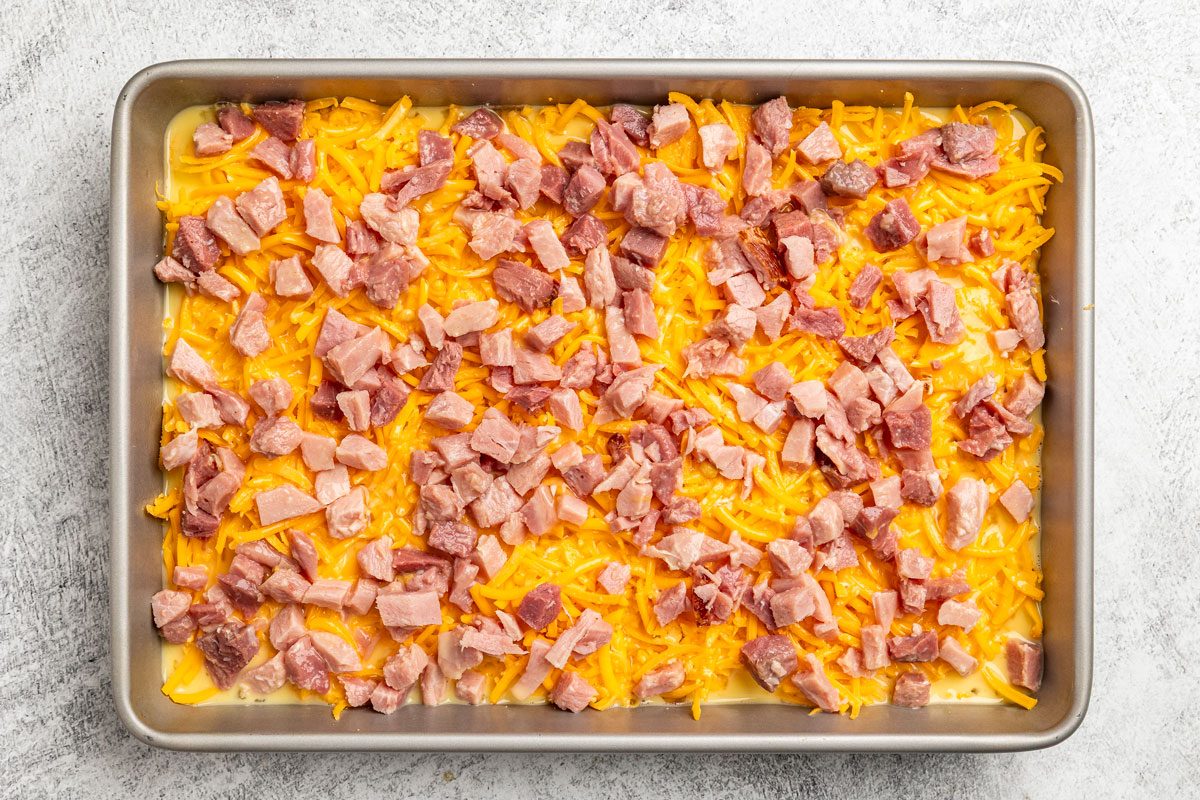 overhead shot of a metal baking tray filled with a layer of Hash Brown Patty Casserole and diced cooked ham on a light colored surface