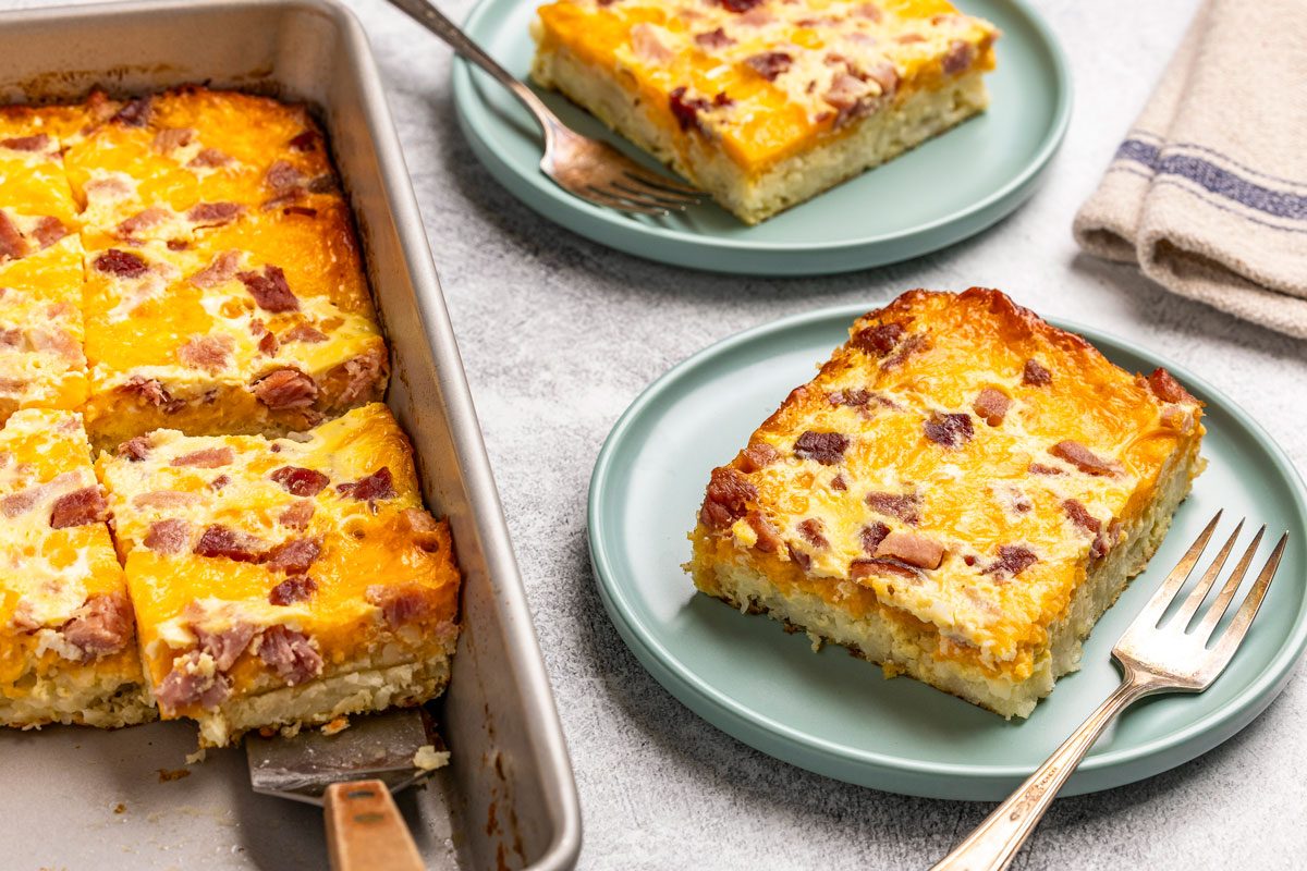 3/4th shot of a baking pan with Hash Brown Patty casserole cut into squares, Two servings are plated with forks on green plates, showing layers of eggs, cheese, and sausage or ham, A beige napkin is nearby