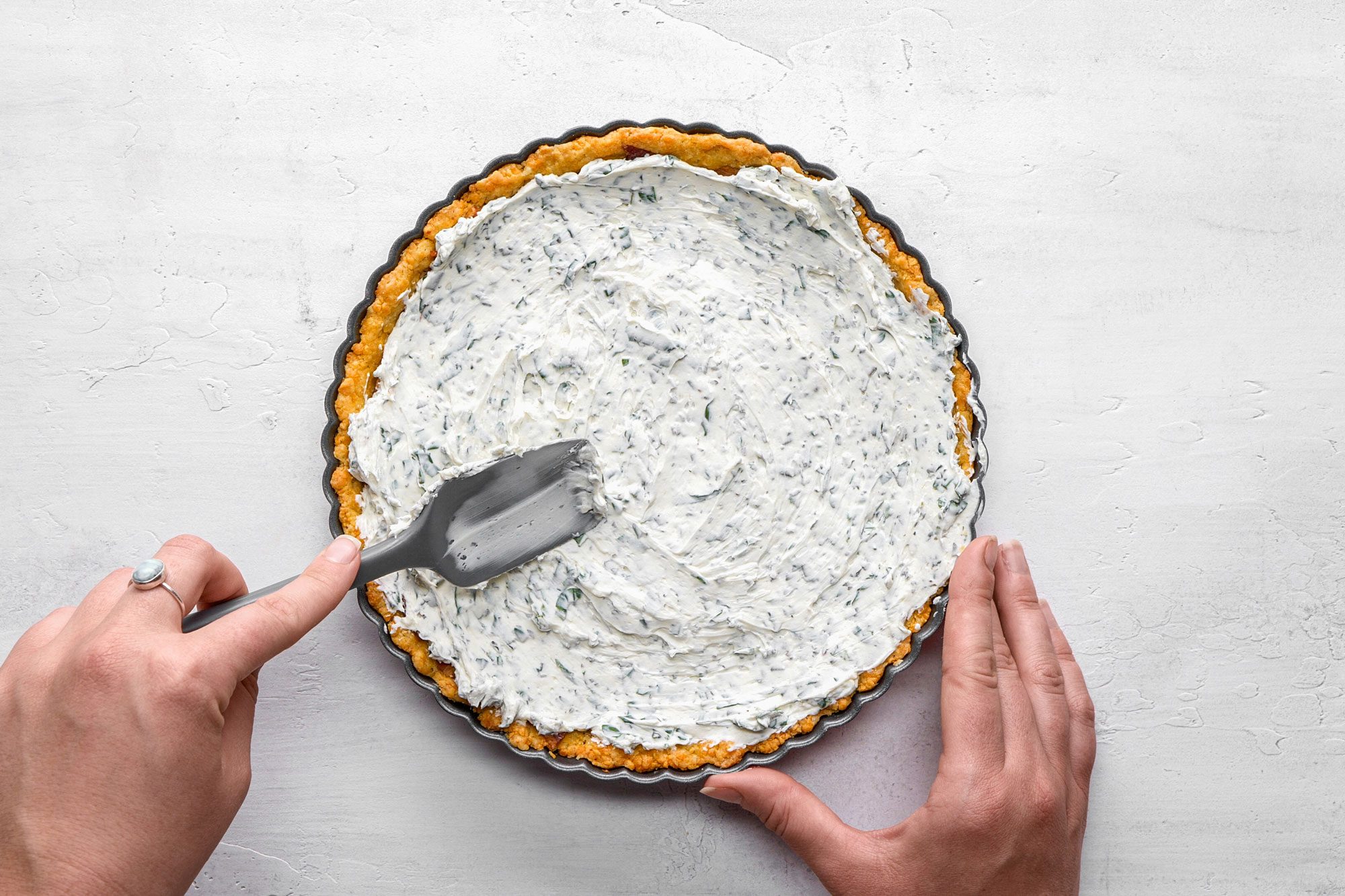 A Top Shot of a person spreads a creamy; herb-filled mixture onto a tart crust in a metal pan using a spatula; steadying the pan on a white surface. The scene suggests baking or preparing a savory tart