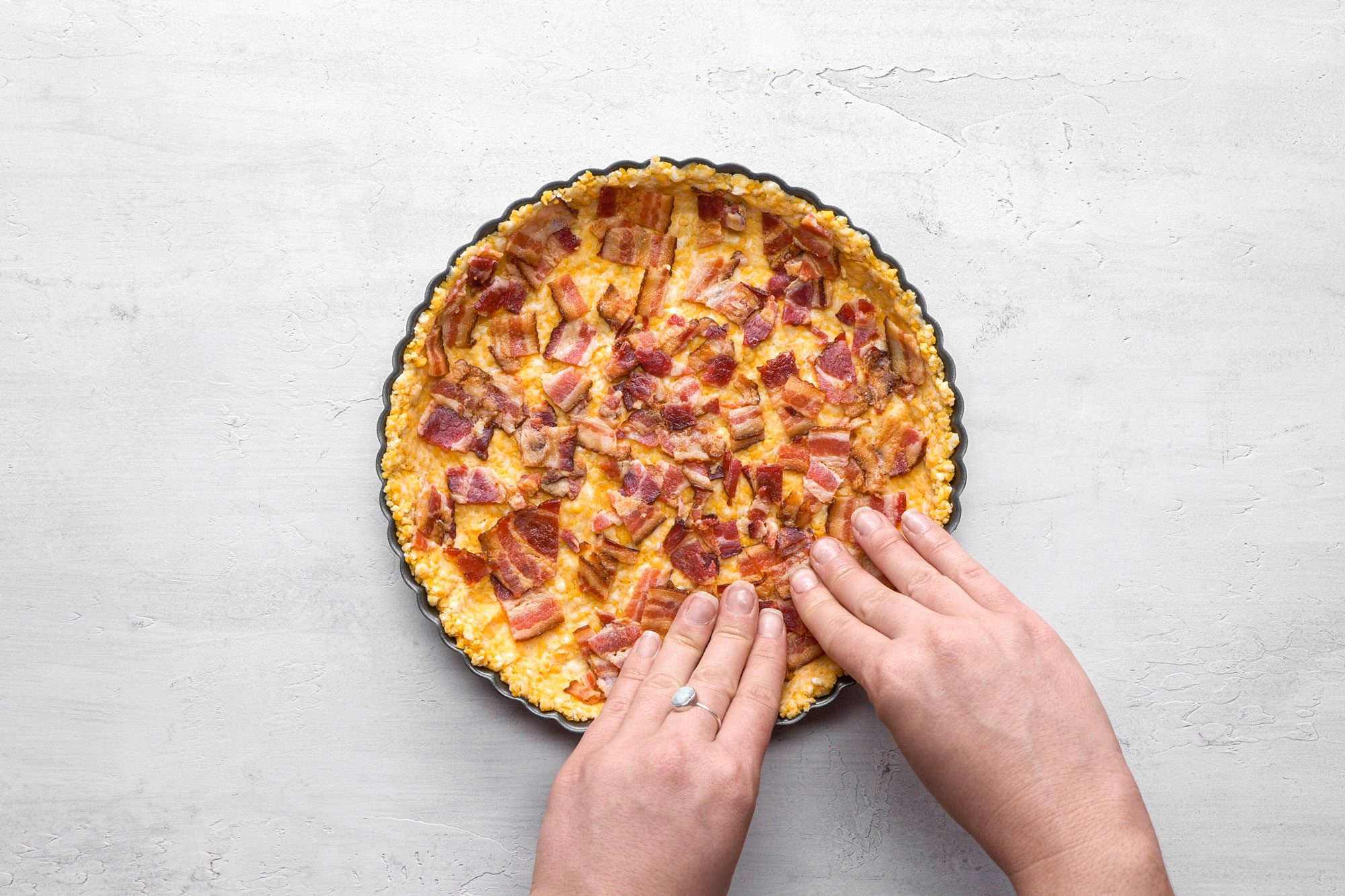A Top shot of a person’s hands hold a freshly baked round quiche with bacon pieces in a baking tin; the scene is set on a light gray textured background, highlighting the golden crust and savory toppings beautifully