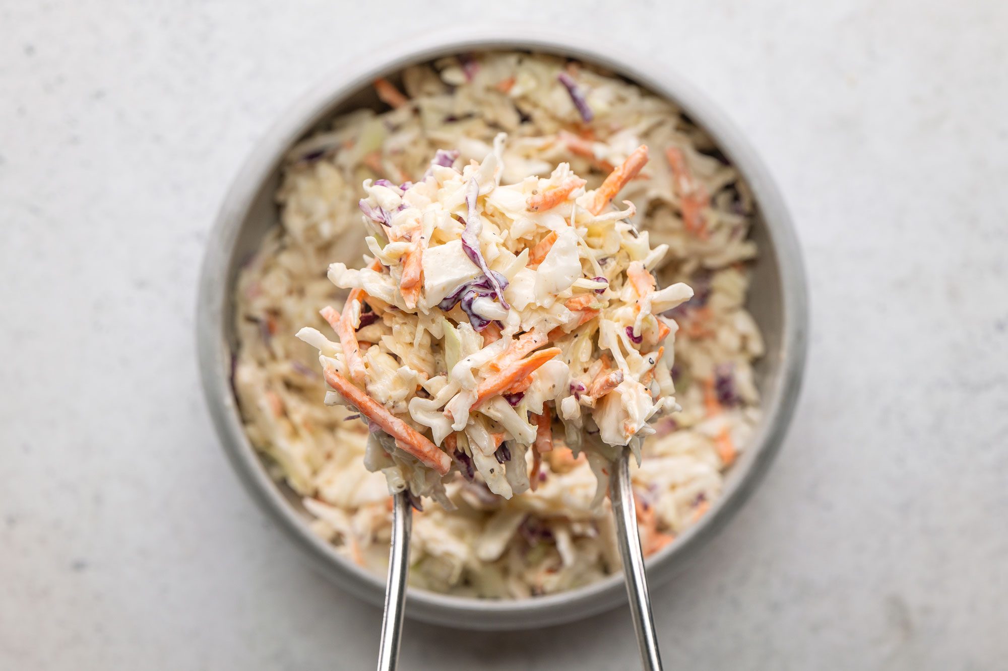 A top shot of a metal tongs lifting creamy coleslaw made of shredded cabbage and carrots from a bowl; viewed from above on a light surface; highlighting freshness and texture