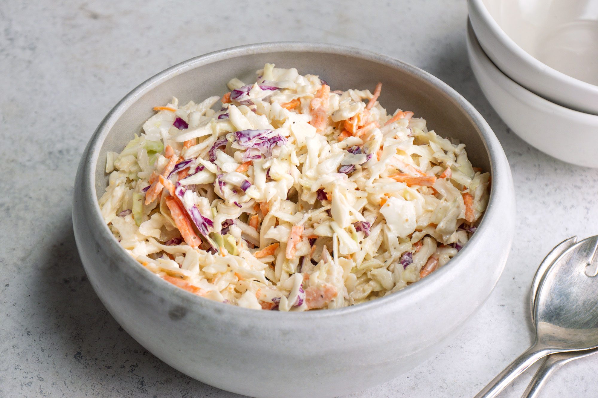 A 3/4th shot of a bowl of creamy coleslaw; featuring shredded cabbage; carrots; and purple cabbage on a light background; accompanied by stacked white bowls and two metal spoons
