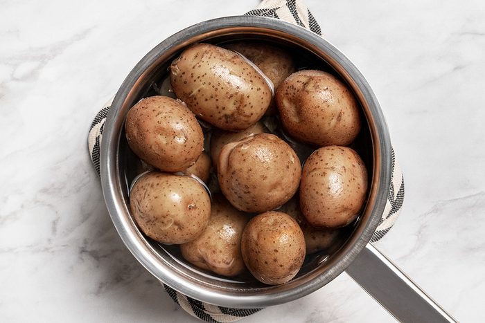 A saucepan filled with unpeeled, brown potatoes sits on a patterned pot holder on a white marble surface.
