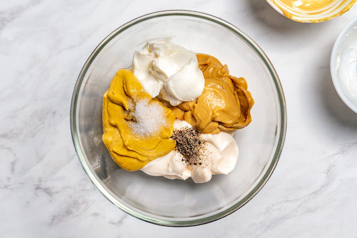 A glass bowl contains dollops of mustard, mayonnaise, sour cream, and spices on a marble surface, ready to be mixed.