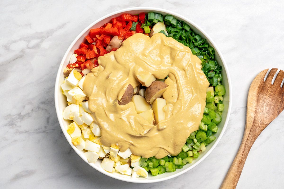A white bowl filled with chopped red bell pepper, green onions, celery, hard-boiled eggs, and potatoes, topped with a generous amount of creamy yellow dressing, sits on a marble surface beside a wooden spoon.