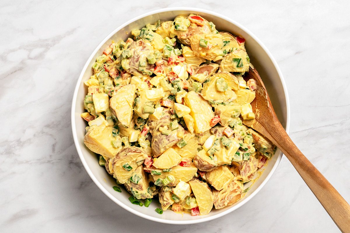 A white bowl filled with creamy potato salad, featuring chunks of potato, chopped vegetables, and herbs, with a wooden spoon resting inside. The bowl sits on a light marble surface.