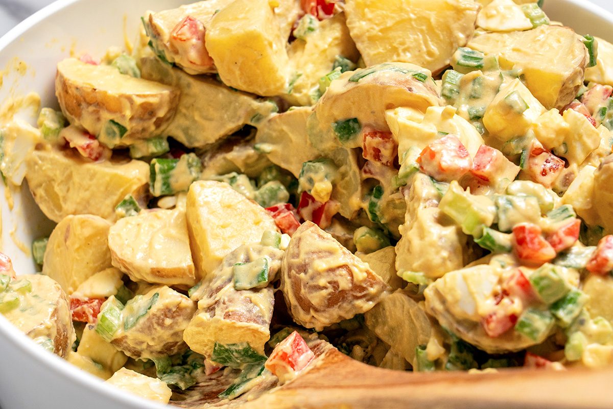 Close-up of a bowl of creamy potato salad with chopped red peppers, green onions, and celery, all mixed together and served with a wooden spoon.
