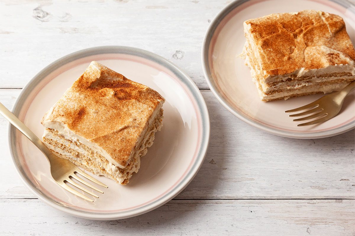 Two plates each hold a square slice of layered dessert topped with cinnamon, next to a gold fork, on a white wooden surface.