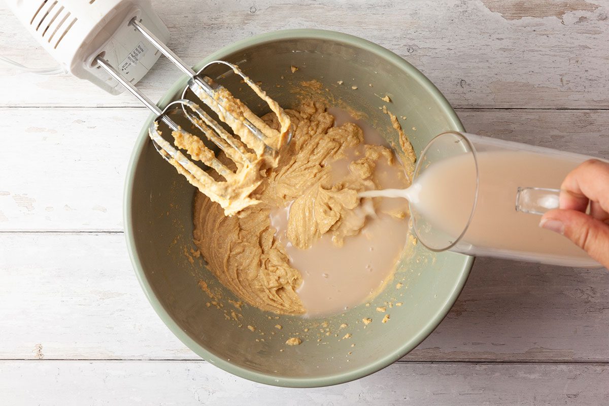 A person pours a glass of milk into a mixing bowl containing batter while using an electric hand mixer, on a light wooden surface.