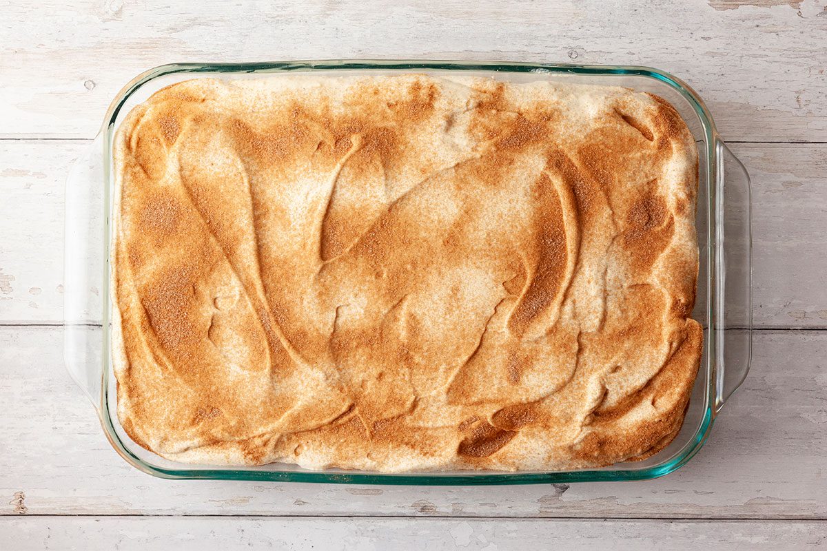 A rectangular glass baking dish filled with a golden-brown baked dessert, possibly cobbler or cake, sits on a light wooden surface. The top has a textured, lightly browned crust.