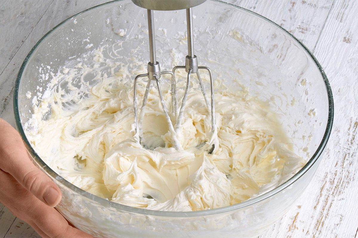 A hand holds a glass bowl filled with whipped cream cheese mixture being blended by an electric hand mixer on a white wooden surface.