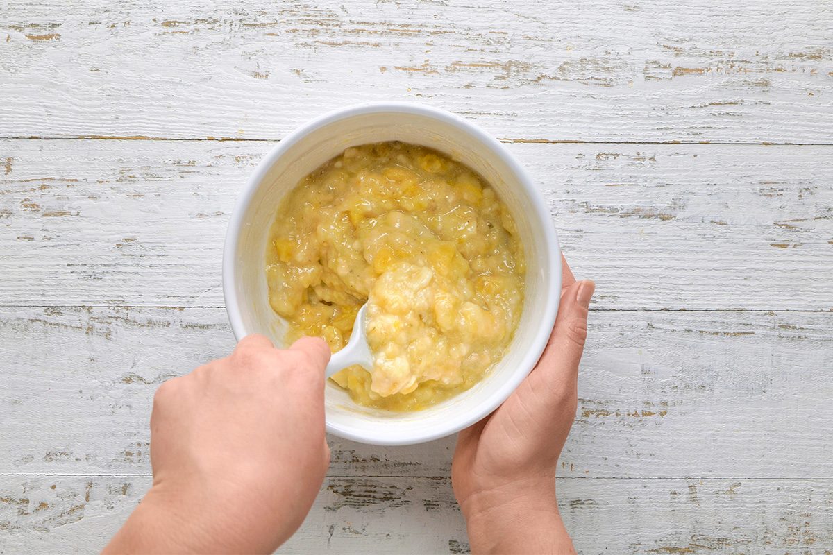 Two hands hold a white bowl of mashed bananas, with a fork mashing the fruit, on a light wooden surface.