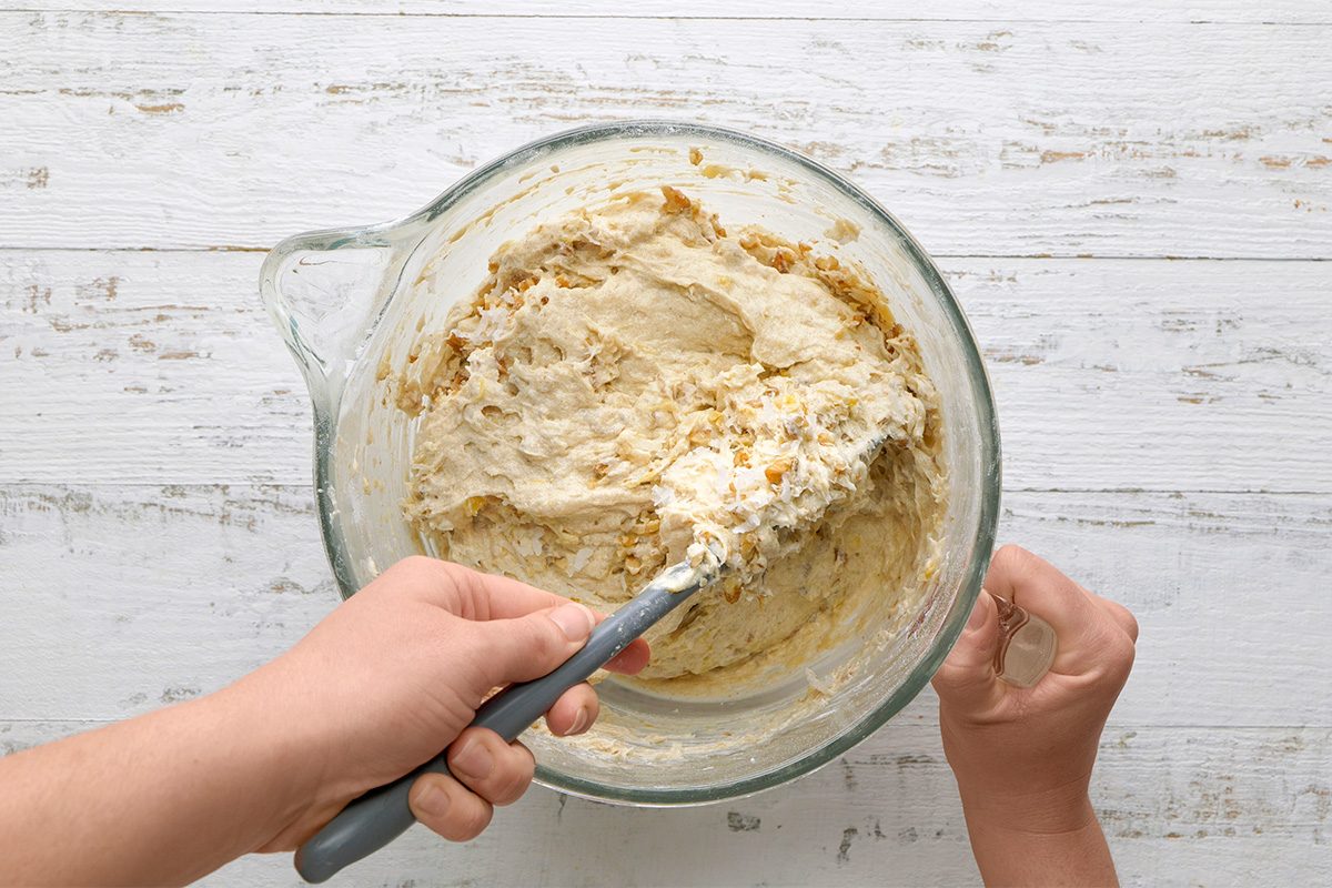 A person mixes dough with a spatula in a clear glass bowl on a white wooden surface.