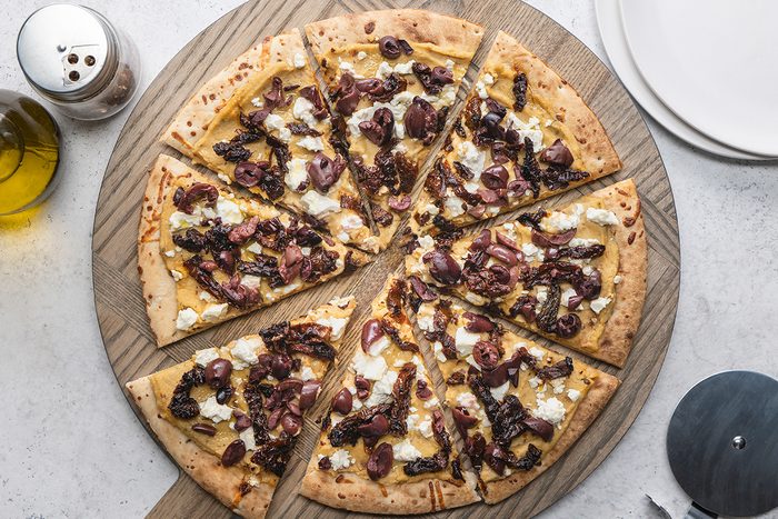 A sliced pizza topped with cheese, caramelized onions, and olives on a round wooden board, with a bottle of olive oil, a pepper shaker, and empty plates nearby.