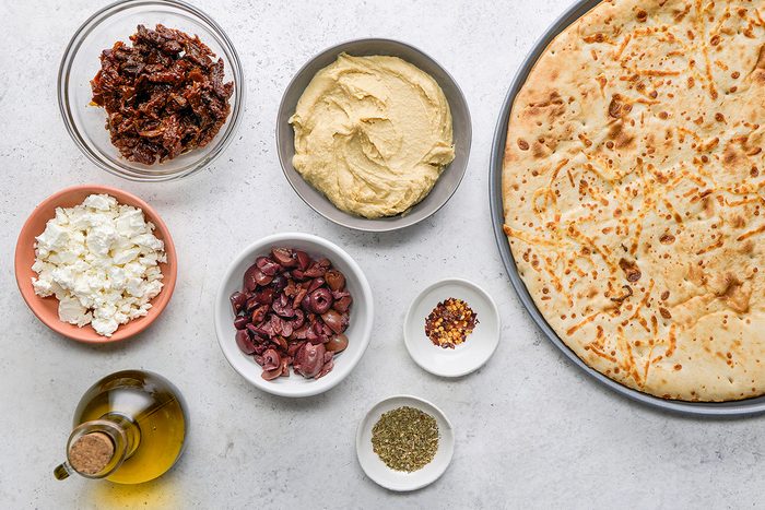 A flatbread sits on a tray next to bowls of hummus, sun-dried tomatoes, feta cheese, kalamata olives, olive oil, crushed red pepper flakes, and dried oregano on a white surface.