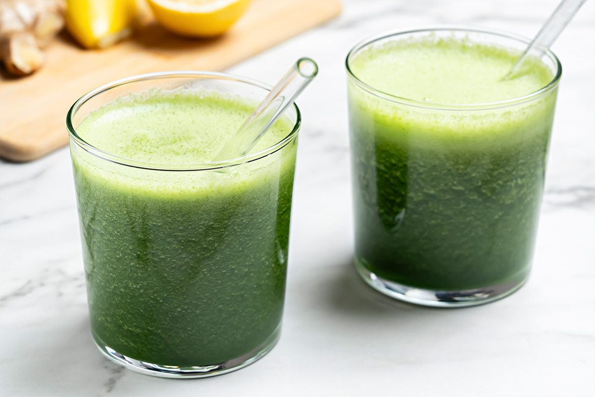 Two clear glasses filled with a frothy green juice, each with a glass straw, are placed on a white marble surface. In the background, there are lemons and ginger on a wooden board.