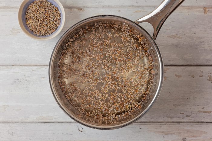 Top shot of a saucepan with water and lavender buds sits on a rustic white wooden surface; beside it is a small bowl of dried lavender buds; creating a calming herbal scene