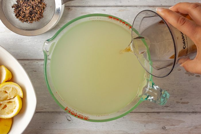 Top Shot of a person pours liquid from a measuring cup into a glass jug of cloudy yellow lemonade; sliced lemons sit in a bowl; tea leaves rest in a strainer on a white wooden surface