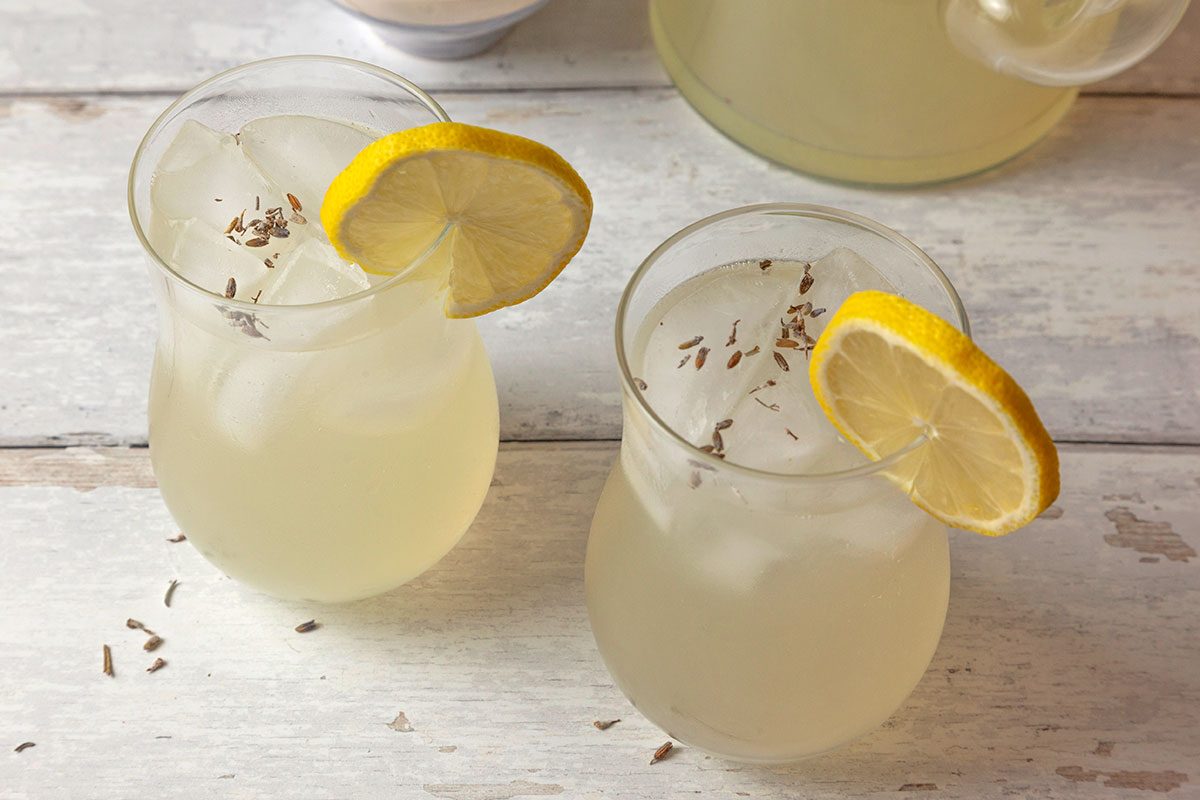 3/4th Shot of a Two glasses of lemonade with ice; lemon slices and lavender buds garnish them; beside a pitcher on a rustic white wooden surface