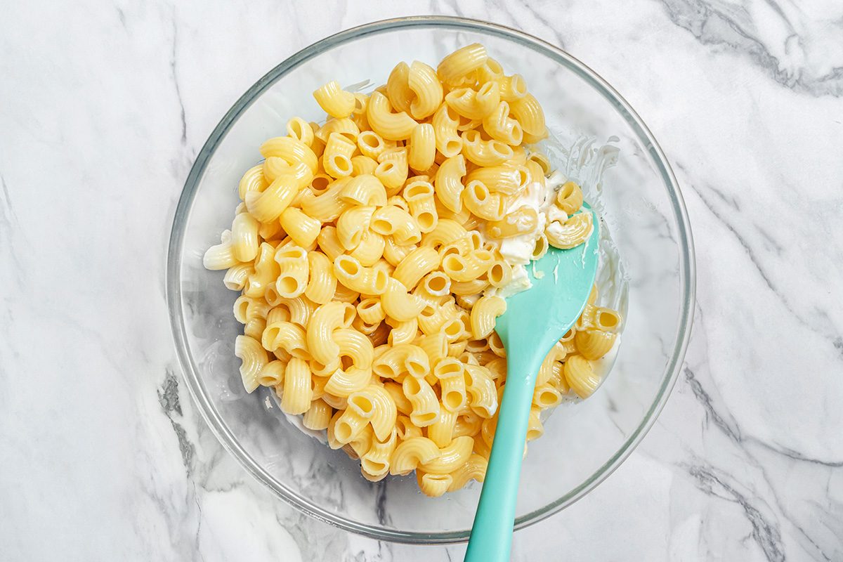 A glass bowl filled with cooked elbow macaroni and shell pasta sits on a marble surface. A turquoise spatula is resting in the bowl, partially mixing the pasta.