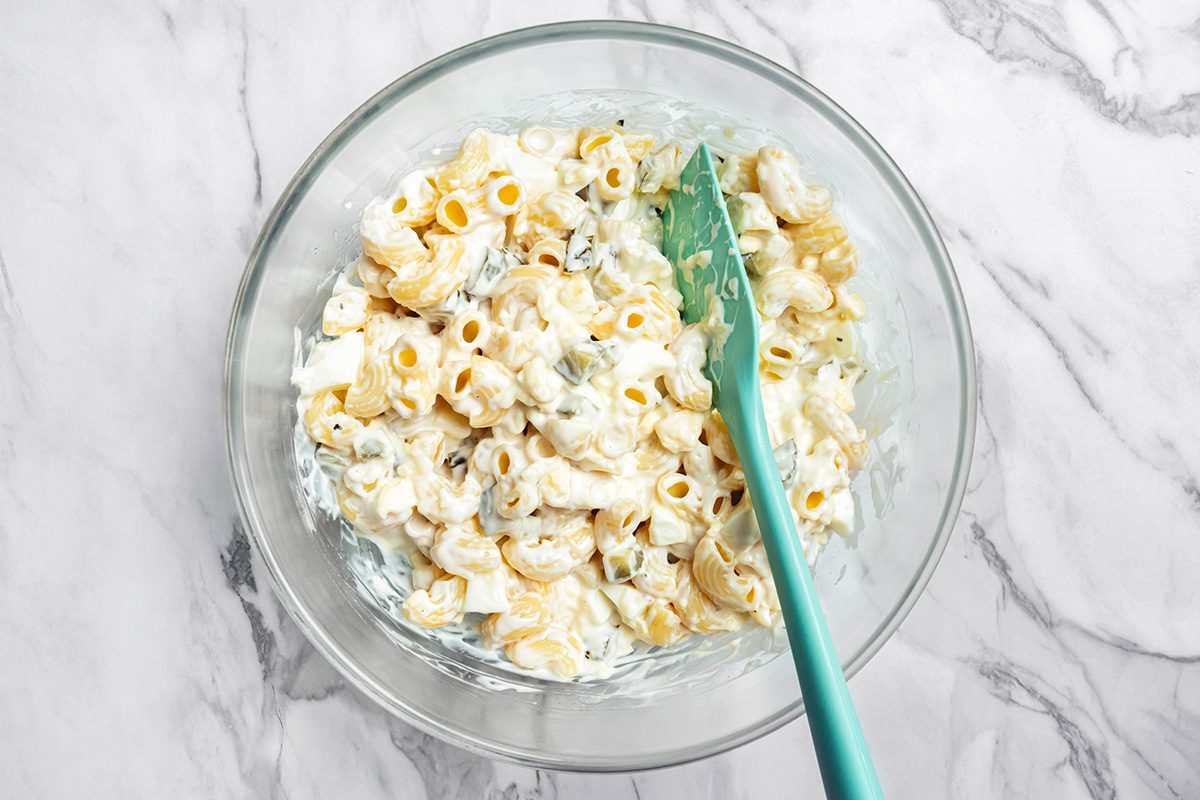 A glass bowl filled with macaroni salad mixed with a creamy dressing, with a teal spatula resting inside. The bowl is placed on a marble surface.