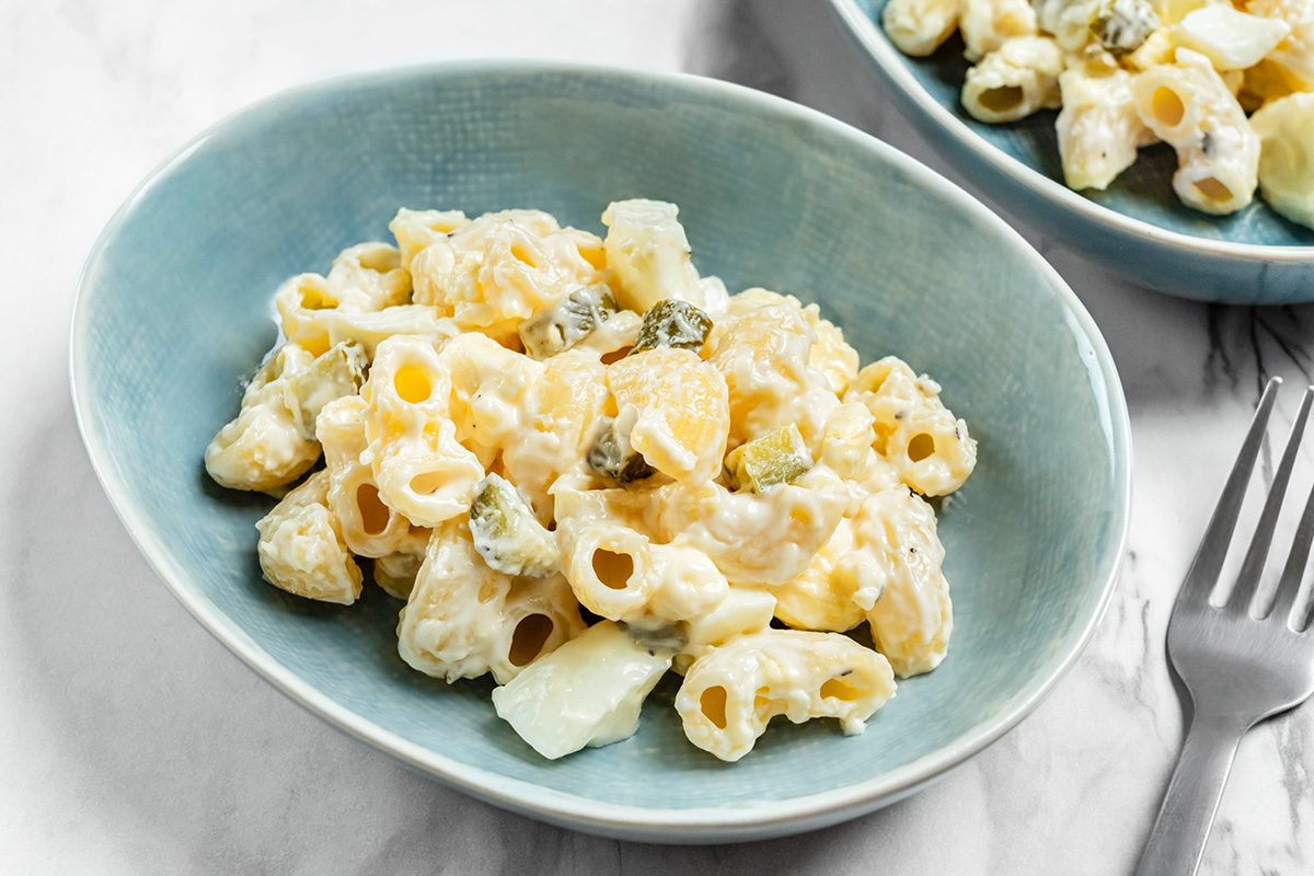 A blue bowl filled with creamy macaroni salad, featuring pasta, chopped pickles, and diced potatoes, sits on a white surface next to a silver fork and another bowl of salad in the background.