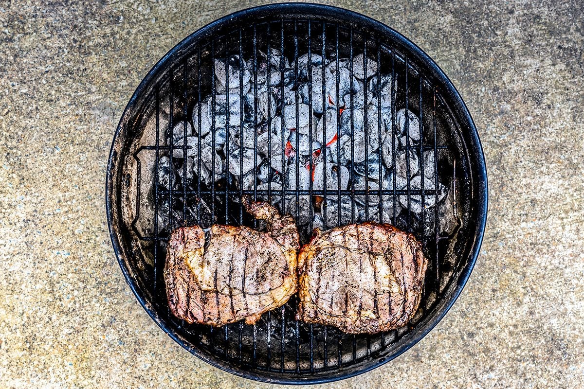 Two seasoned steaks sitting over coals on a small outdoor grill